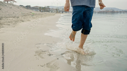 A young boy running barefoot on the sandy beach and enjoying the cool sea water. Fun, happiness, and excitement of a family vacation, journey, and weekend. © Кирилл Рыжов