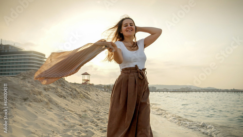 Young woman twirls a colorful silk scarf around at the beach as she smiles and laughs in the wind. Emphasis on happiness  travel  and leisure.