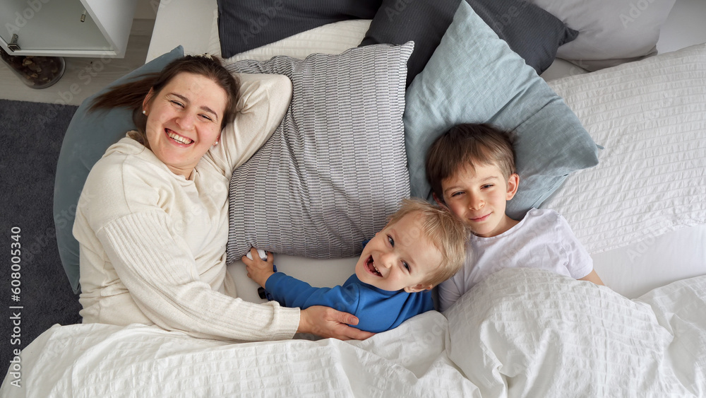 Happy cheerful mother with two sons lying in bed at morning and smiling in camera. Concept of family happiness, relaxing at home, having fun in bed, parent and cheerful kids