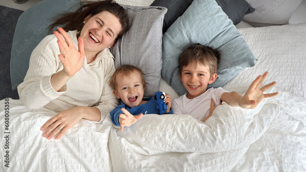 Young happy mother lying in bed with her two sons and waving in camera. Concept of family happiness, relaxing at home, having fun in bed, parent and cheerful kids.