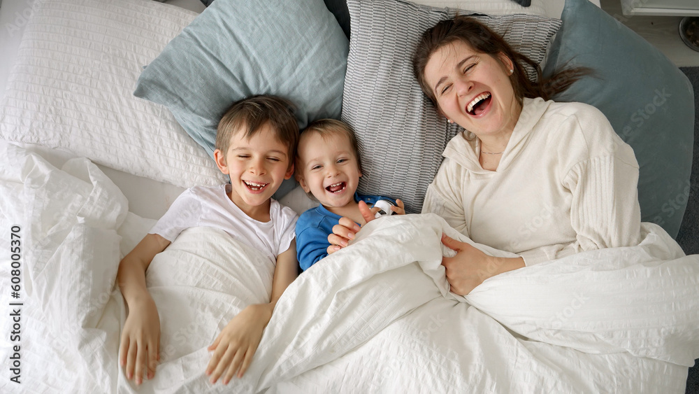 A cheerful young mother in bed with her two sons waving at the camera. This captures the essence of family happiness, spending quality time at home, and joyous moments between parent and child. A cheerful young mother in bed with her two sons waving at the camera. This captures the essence of family happiness, spending quality time at home, and joyous moments between parent and child.