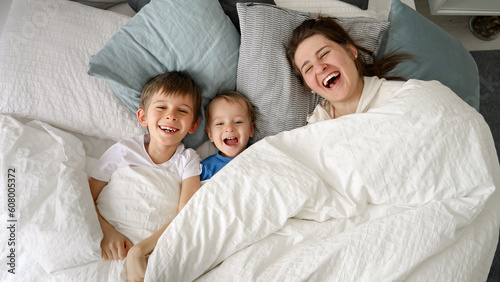 Happy laughing family with two kids hiding under blanket in bed and throwing it off. Concept of family happiness, relaxing at home, having fun in bed, parent and cheerful kids