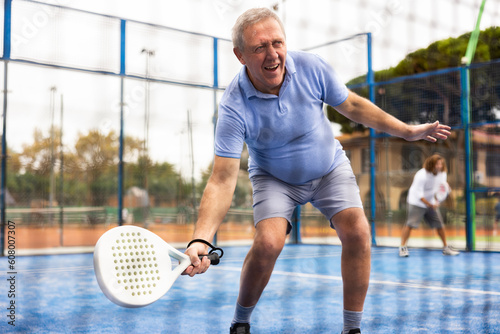 Positive elderly male player serving ball during training padel in court. View through tennis net
