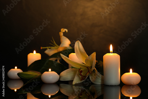 White lilies and burning candles on black mirror surface in darkness, space for text. Funeral symbols