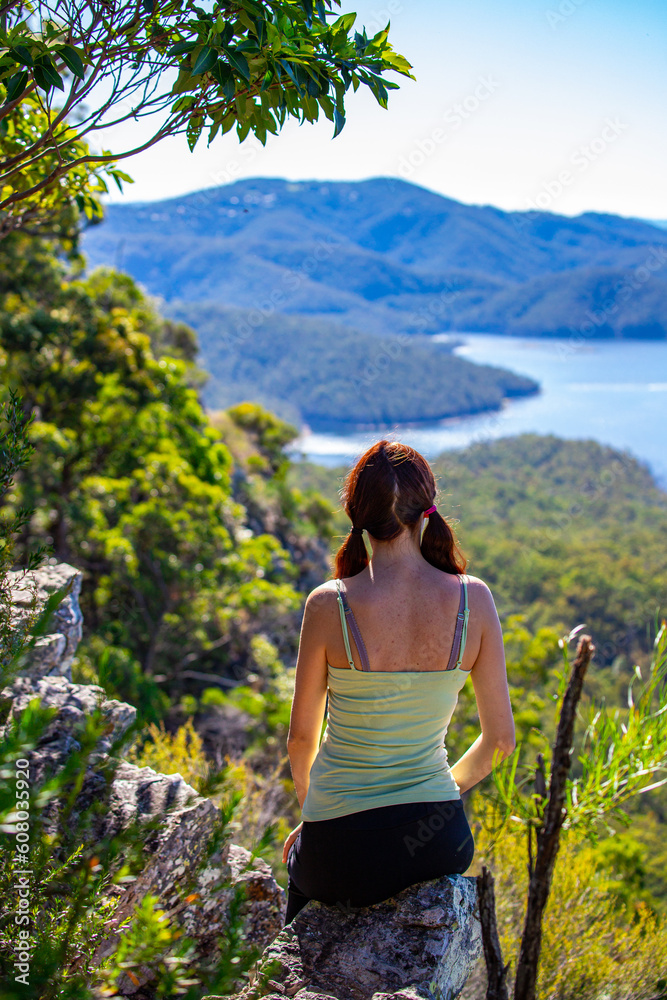 Naklejka premium Beautiful girl sits on a rock shelf enjoying view of the Advancetown lake after successfully climbing the Pages Pinnacle. Hidden gems in Springbrook National Park, Gold Coast, Queensland, Australia