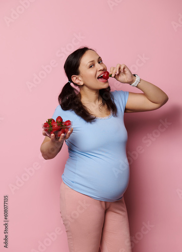 Gorgeous pregnant woman eating fresh organic strawberries, isolated over pink background. Expectant mother in blue t-shirt, holding bowl with fresh berries. Healthy nutrition and pregnancy concept.