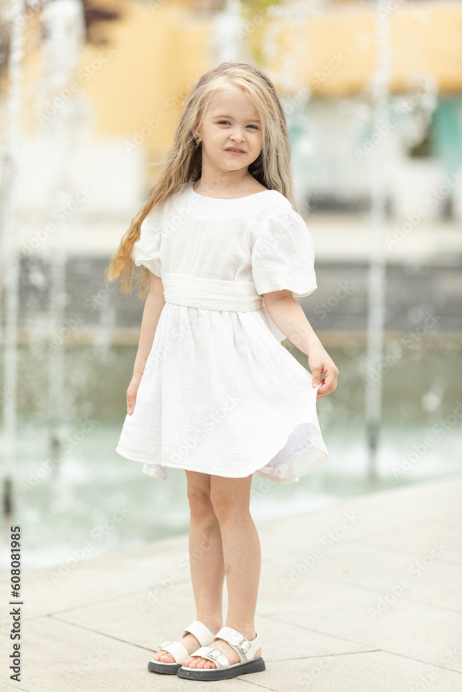 Summer portrait of cute little blonde girl in white dress against the backdrop of a fountain. space for text. summer concept. children's day theme.