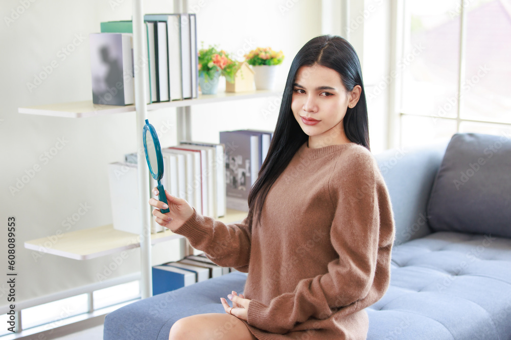 Millennial Asian beautiful pretty female model in casual outfit big brown warm sweater sitting on cozy sofa couch holding handheld mirror posing in living room. Millennial Asian beautiful pretty female model in casual outfit big brown warm sweater sitting on cozy sofa couch holding handheld mirror posing in living room.