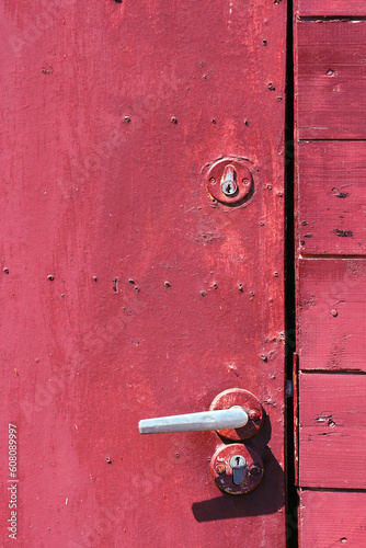 Fragment of a closed old door sloppily painted crimson with an aluminum door handle. Red background or texture. Vertical photo.