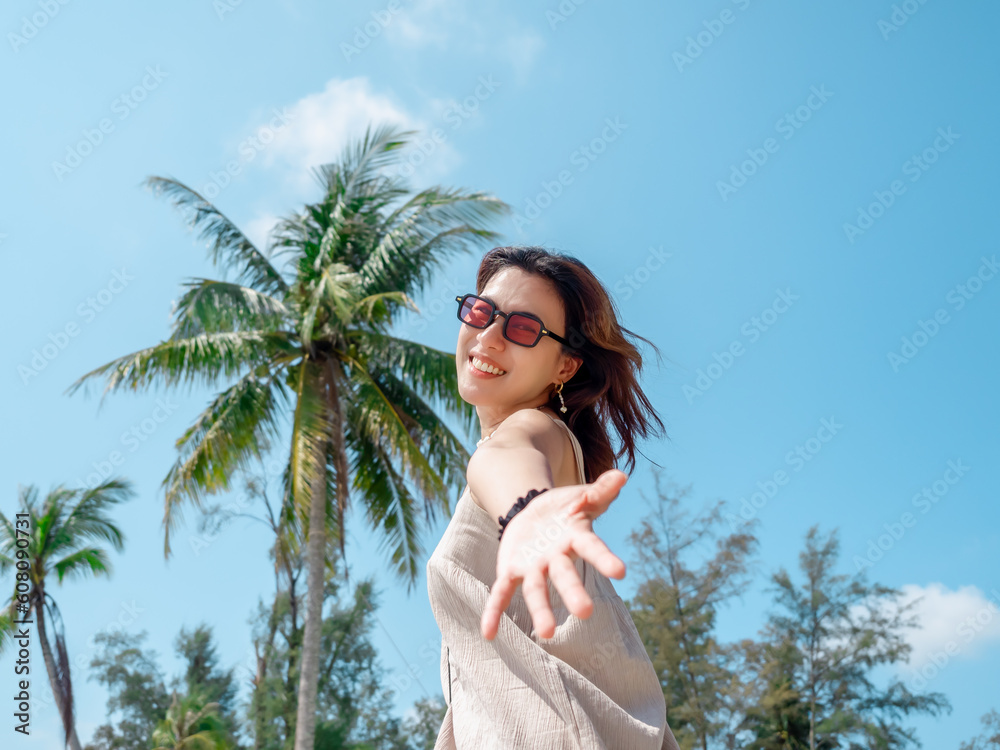 Beautiful Asian woman portrait with sunglasses on the beach in summer vibes. Happy female smile looking at camera in front of tropical palm trees on blue sky background, Holiday vacation, summertime.