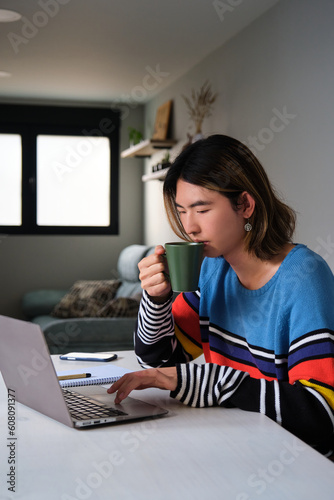 Young taiwanese man drinking coffee while working at home with a laptop.