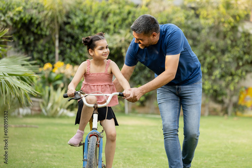 Fototapet Indian father teaching riding a cycle to his daughter in lawn
