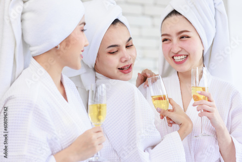 Three Asian cheerful happy beautiful female girlfriends in casual bathrobe and towel outfit standing smiling holding sparkling champagne tall glass cheers toasting together in spa massage studio