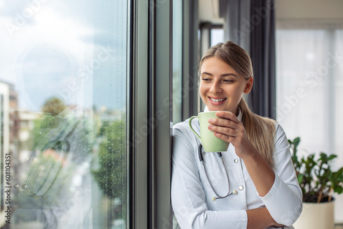 Shot of female doctor holding cup of coffee while looking forward window standing in the consultation. Smiling young woman doctor in white medical uniform and stethoscope look in distance planning.