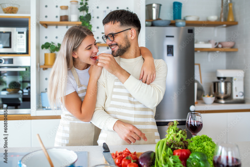 Beautiful young couple is looking at each other and feeding each other with smiles while cooking in kitchen at home. Loving joyful young couple embracing and cooking together