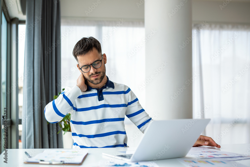 Businessman feeling pain in neck after sitting at the table with laptop. Tired man suffering of office syndrome because of long hours computer work. He is massaging his tense neck muscles Businessman feeling pain in neck after sitting at the table with laptop. Tired man suffering of office syndrome because of long hours computer work. He is massaging his tense neck muscles