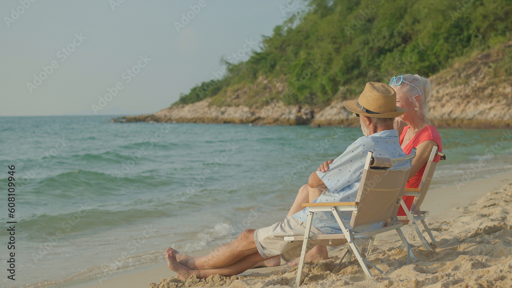 A couple of elderly sitting in chairs at the beach watching the sun and the sea on their summer vacation and they smile and enjoy their vacation.