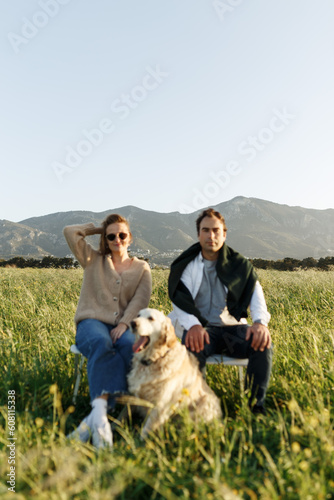 Young happy couple with dog, sitting on chairs in feather grass field on summer sunset day, looking forward in camera, serious.
