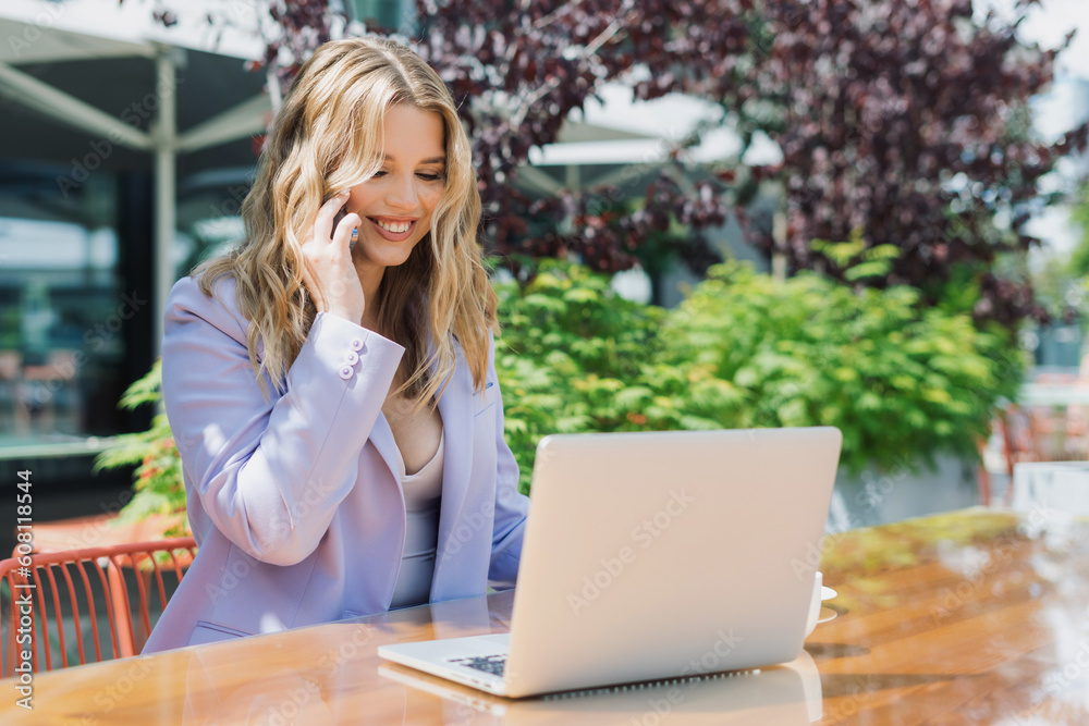 A beautiful blonde in a stylish suit is talking on the phone sitting in front of a laptop. Work on the summer terrace A beautiful blonde in a stylish suit is talking on the phone sitting in front of a laptop. Work on the summer terrace