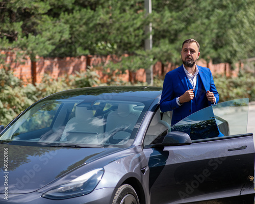 Caucasian bearded man in a blue suit gets out of a black electro car in the countryside in summer.