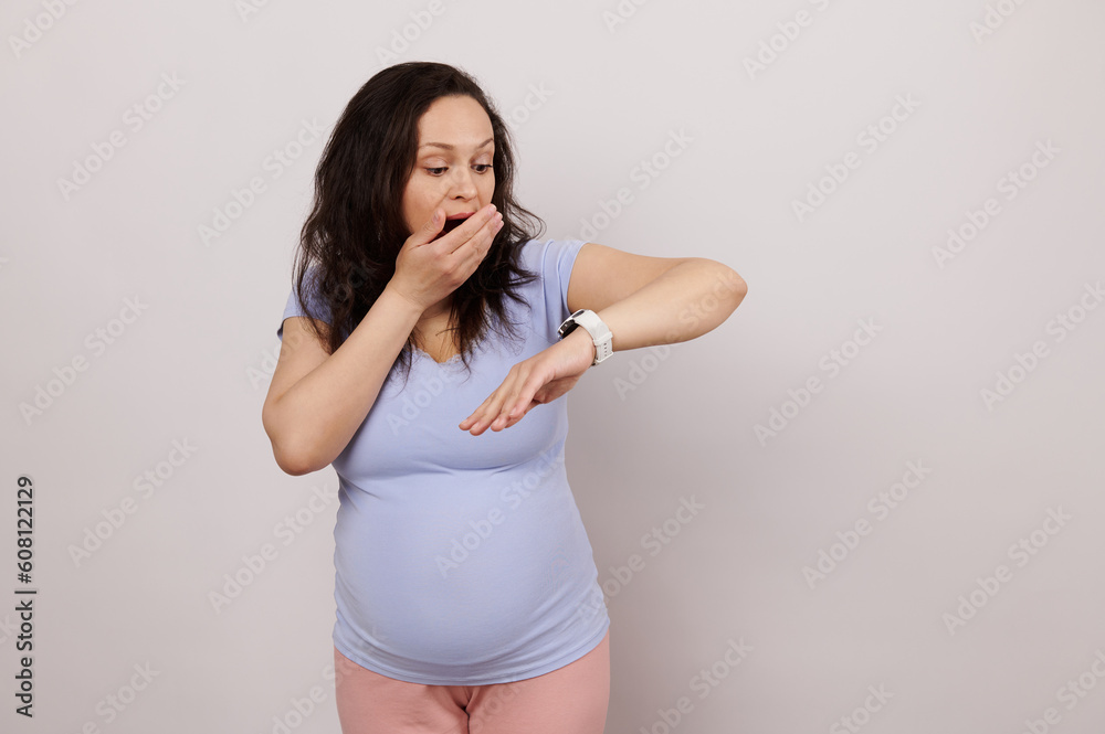 Young amazed pregnant woman looking at the monitor of her electronic smart wrist watch, expressing stupefaction, checking time, using health monitor app, internet, messaging, isolated white background Young amazed pregnant woman looking at the monitor of her electronic smart wrist watch, expressing stupefaction, checking time, using health monitor app, internet, messaging, isolated white background