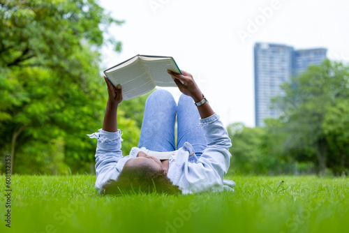 African American woman is lying down in the grass lawn inside the public park holding book in her hand during summer for reading and education concept