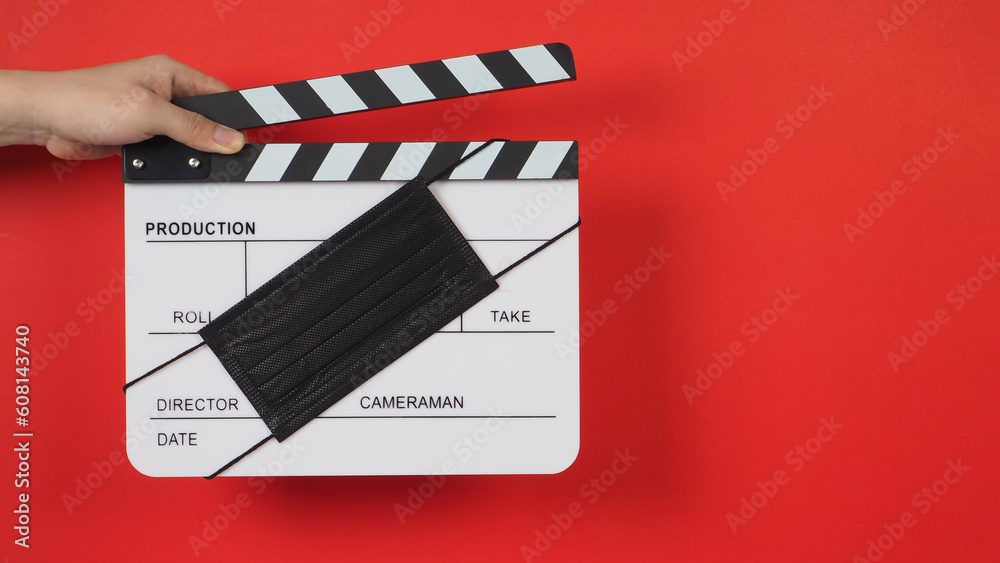 Hand is holding white clapper board with a black face mask on red background.