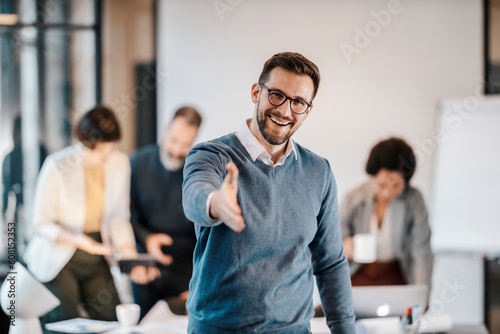 Portrait of a happy executive introducing himself while standing at the office with his colleagues in blurry background.