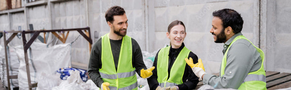 Cheerful multiethnic sorters in high visibility vests and gloves talking while standing near trash in outdoor waste disposal station, garbage sorting and recycling concept, banner Cheerful multiethnic sorters in high visibility vests and gloves talking while standing near trash in outdoor waste disposal station, garbage sorting and recycling concept, banner