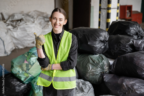 Smiling young worker in protective vest and gloves showing like gesture and looking at camera while standing near blurred plastic bags with trash in garbage sorting center, recycling concept