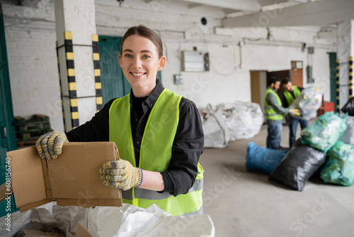 Cheerful young worker in protective vest and gloves holding cardboard and looking at camera while working near sack and blurred colleagues in garbage sorting center, recycling concept