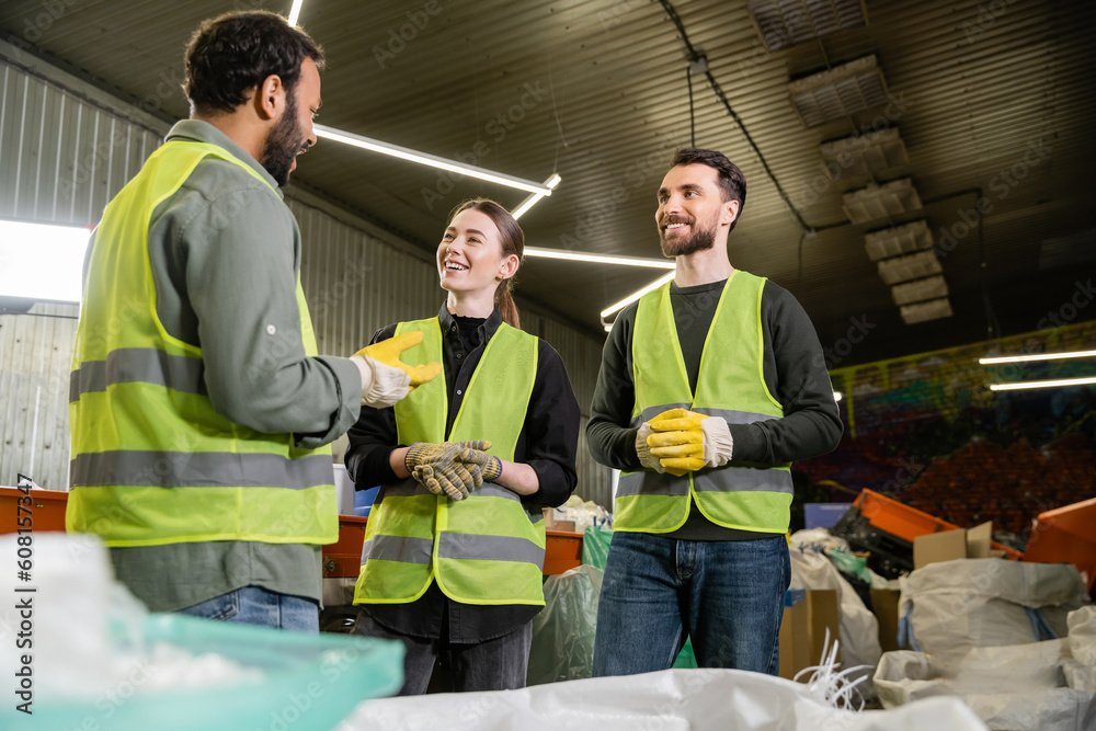 Bearded indian worker in protective glove and vest talking on cheerful colleagues while standing together near blurred sacks with trash in waste disposal station, recycling concept Bearded indian worker in protective glove and vest talking on cheerful colleagues while standing together near blurred sacks with trash in waste disposal station, recycling concept