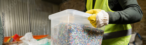 Cropped view of sorter in protective vest and glove holding container with plastic for recycle while working in blurred garbage sorting center, garbage sorting and recycling concept, banner