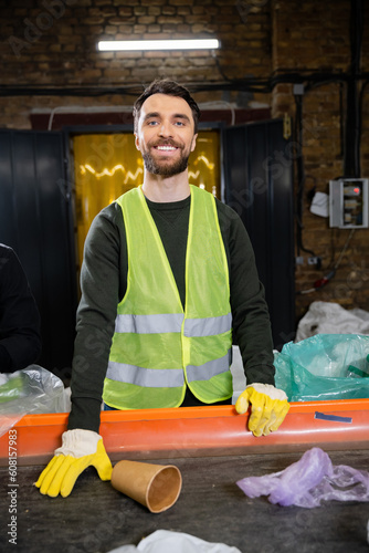 Smiling and bearded sorter in gloves and protective vest looking at camera while working with trash on conveyor in garbage sorting center, garbage sorting and recycling concept