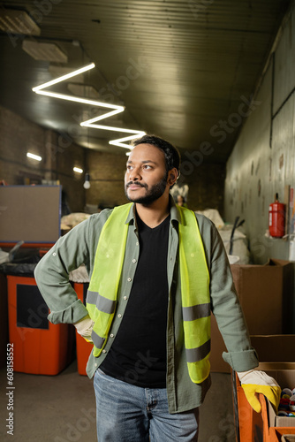 Indian man in high visibility vest and gloves looking away while standing near plastic caps in carton box and working in garbage sorting center, garbage sorting and recycling concept
