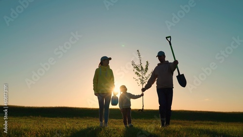 Happy family and child, team planting trees in spring. Silhouette of family with tree. Dad is farmer, mom is child planting trees. Green nature concept. Gardeners with shovel go to plant tree, sunset