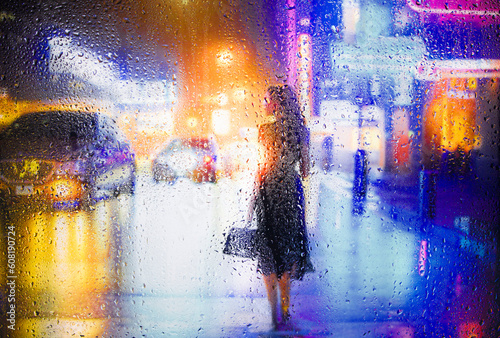 View through glass window with rain drops on blurred reflection silhouette of a girl on a city street after rain and colorful neon bokeh city lights  night street scene. Focus on raindrops on glass
