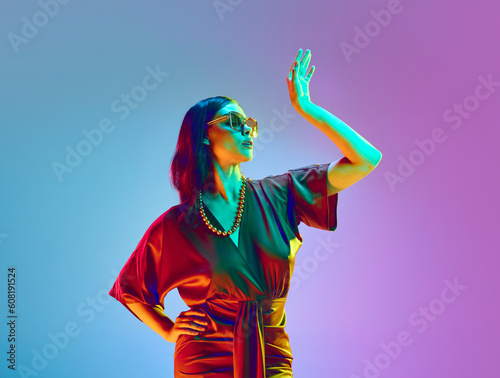 Dreaming girl, fashion model with bright make-up wearing fashionable silk dress and necklace posing over violet neon background. Concept of beauty, fashion week