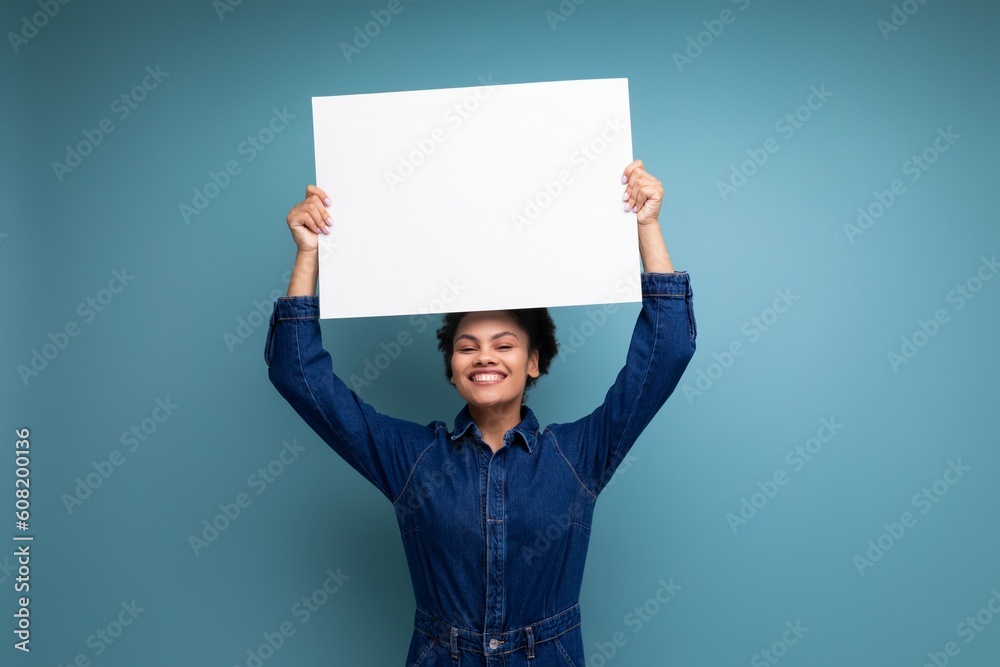 satisfied positive young hispanic brunette woman with fluffy curly hair in blue denim suit holding a white sheet of paper with a mockup for advertising satisfied positive young hispanic brunette woman with fluffy curly hair in blue denim suit holding a white sheet of paper with a mockup for advertising