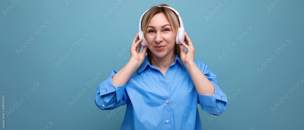 horizontal photo of a smiling young woman in a casual shirt listening to music in big white headphones on a blue isolated background with copy space