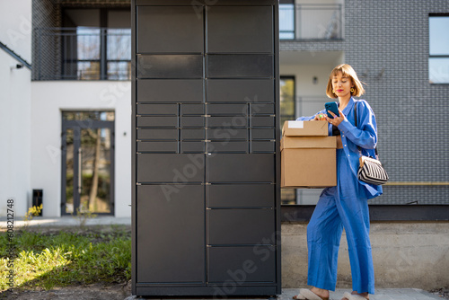 Young woman picks up parcels from automatic post office machine, standing with phone near apartment building. Concept of fast delivery to automatic self lockers photo