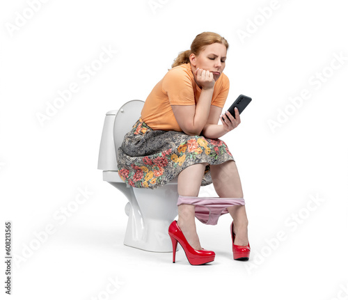 A young woman in a colorful skirt sits on the toilet and looks at her smartphone, isolated on a white background