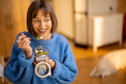 Happy woman enjoys granola breakfast in bowl, standing in modern kitchen at home. Concept of wellness, trendy breakfasts and domestic lifestyle