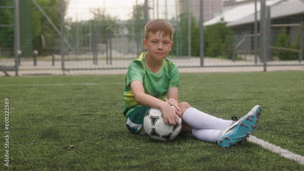 The boy dressed in football uniform is practicing on a mini football stadium. A portrait of a young football player sitting on artificial turf, holding a football in his hands and smiling. The boy dressed in football uniform is practicing on a mini football stadium. A portrait of a young football player sitting on artificial turf, holding a football in his hands and smiling.