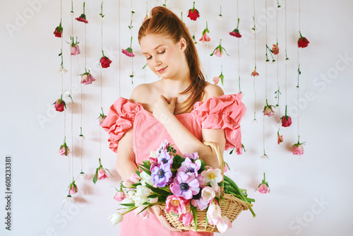 Fototapeta Naklejka Na Ścianę i Meble -  Studio portrait of pretty young teenage 15 - 16 year old red-haired girl wearing pink coral dress, posing on white background with hanging roses, holding basket with many spring flowers