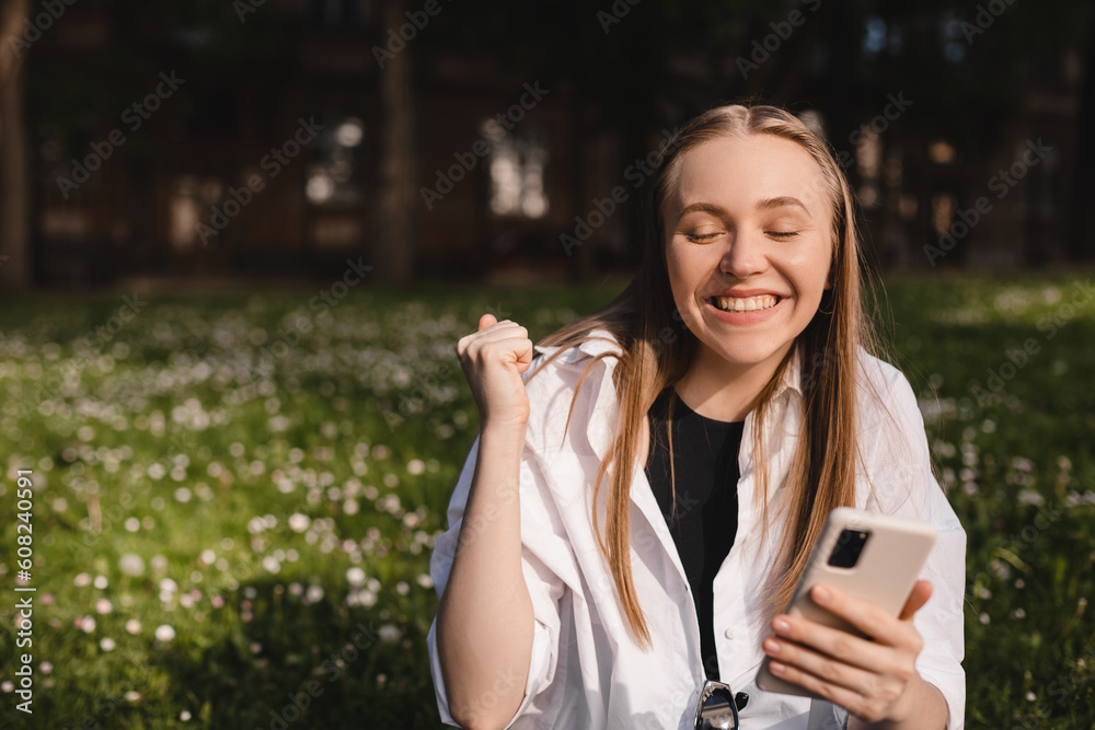 Portrait photo smiling young girl sitting on ground browsing internet with cellphone wearing casual clothes gesturing like winner. Blonde woman look happy, celebrating win, triumph, success. Portrait photo smiling young girl sitting on ground browsing internet with cellphone wearing casual clothes gesturing like winner. Blonde woman look happy, celebrating win, triumph, success.