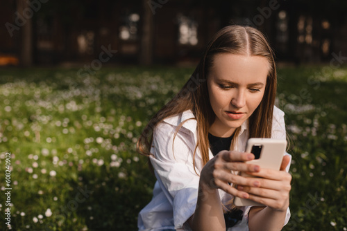 Woman sitting on grass smiling. Pensive businesswoman young caucasian woman student freelancer using smart phone for e-learning, e-banking, mobile applications, surfing internet social media in park.