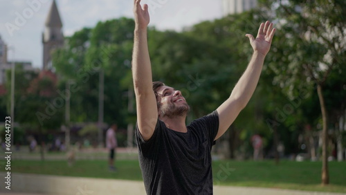 Happy man raising arms to sky thanking God with HOPE and FAITH. A male caucasian person in 30s feeling GRATITUDE standing outdoors gazing upward at park