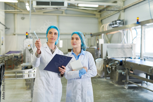 Serious pensive female food production engineer pointing with pen at machine while discussing manufacture with colleague in bakery workshop