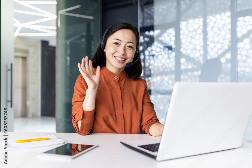 Portrait of successful tech support worker, Asian woman looking at camera, businesswoman with headset for video call, greeting gesture, inside office with laptop. Portrait of successful tech support worker, Asian woman looking at camera, businesswoman with headset for video call, greeting gesture, inside office with laptop.
